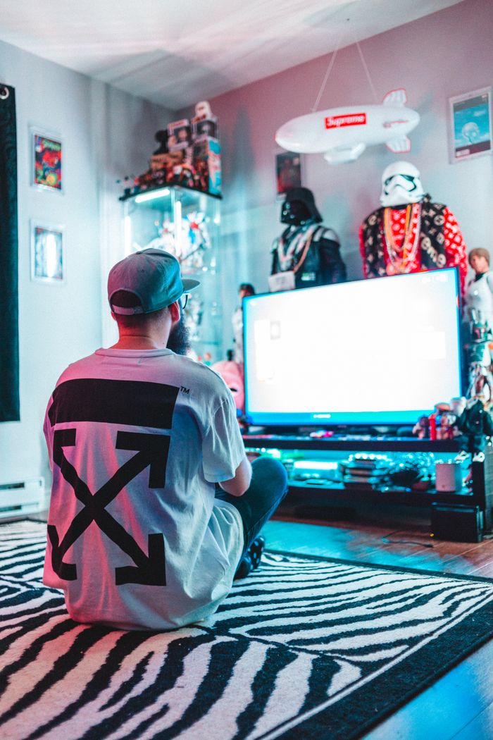 Back view of unrecognizable male in casual clothes and cap sitting on floor near TV and playing video game in room